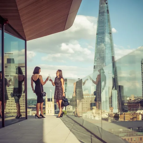 Two women standing on a balcany cheersing, with a London city in the background